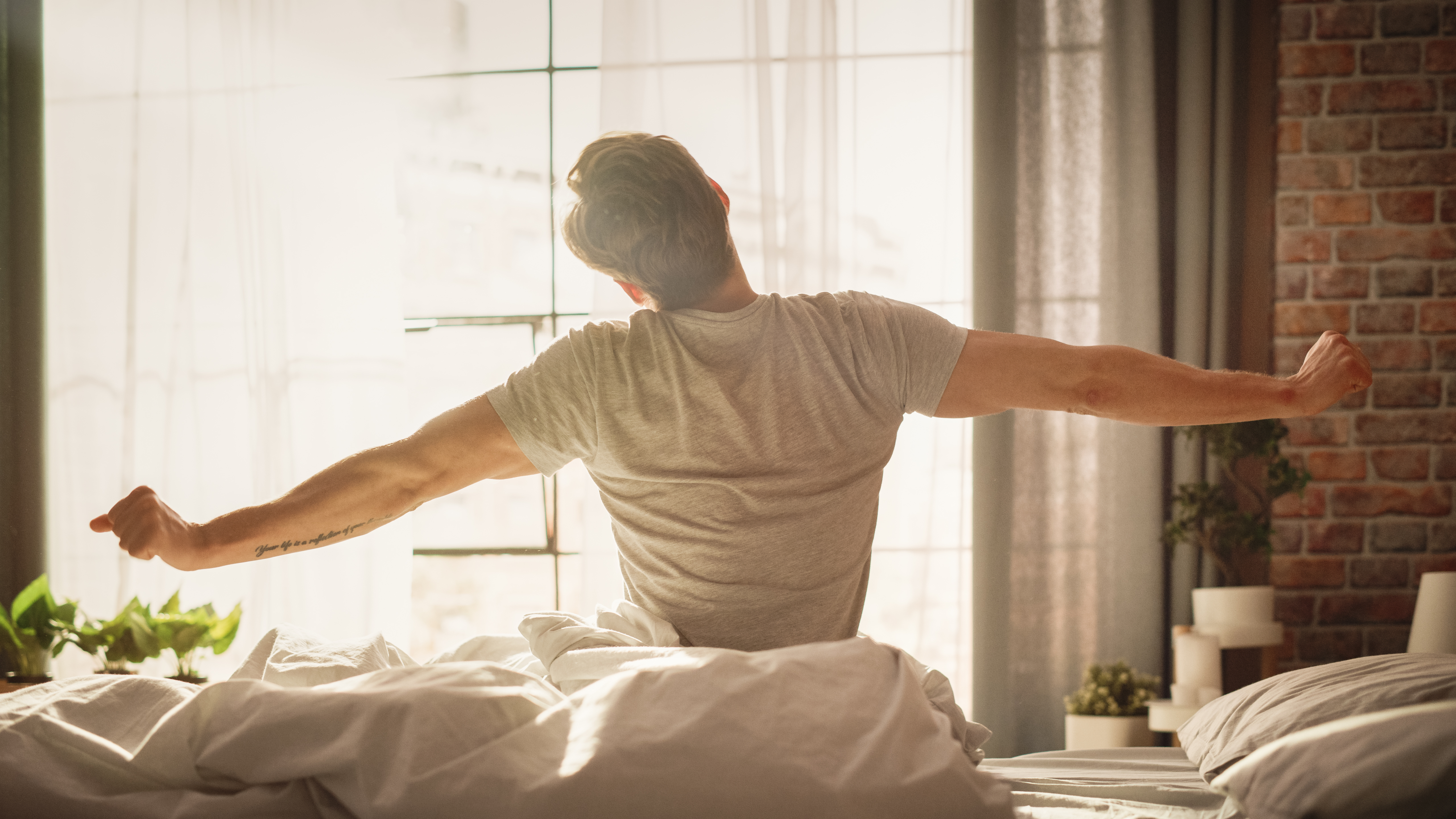 Man stretching in bed, morning light through window, relaxed atmosphere.