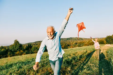 Happy middle-aged couple running and flying a red kite in an open field