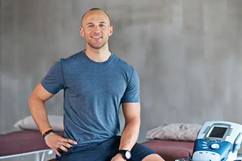 Man in a gym, smiling, sitting beside medical equipment.