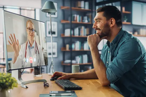Man in a video call with a doctor on a computer, smiling in a modern home office.