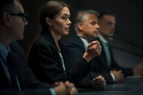 Women and men in suits at a conference table, one speaking into a microphone.