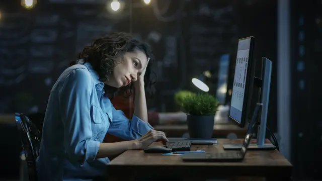 Woman in blue shirt, tired, working on a computer at night.