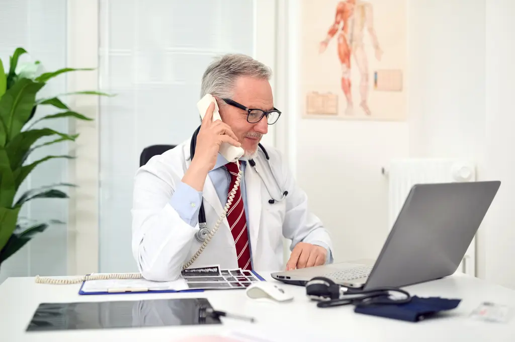 Male doctor talking on phone at desk with laptop open