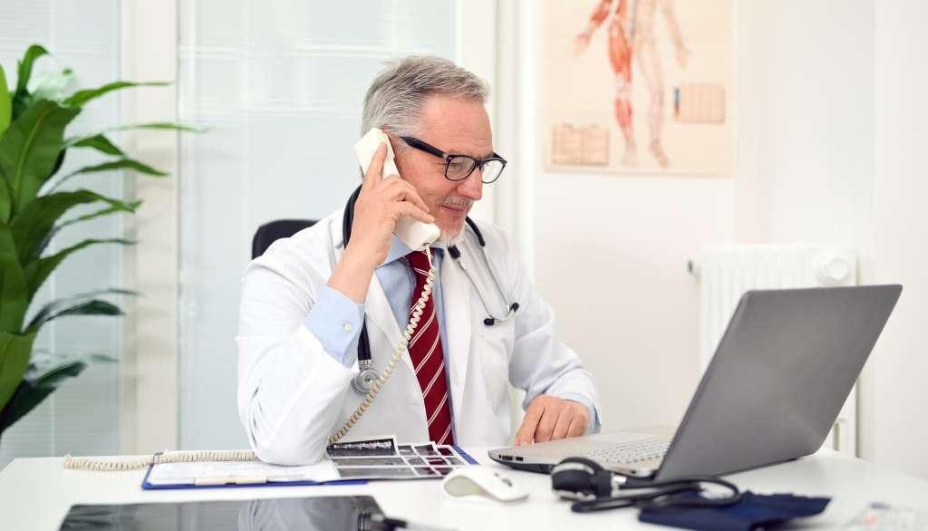 Male doctor talking on phone at desk with laptop open