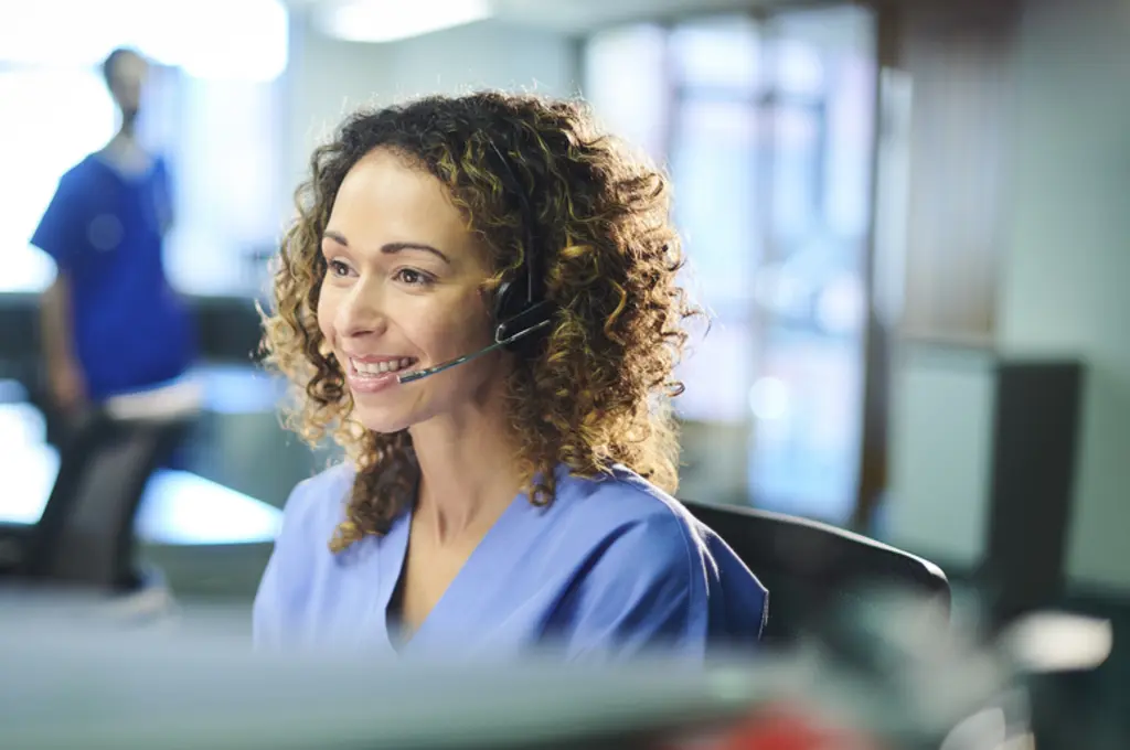 Smiling woman in scrubs with a headset at a desk.