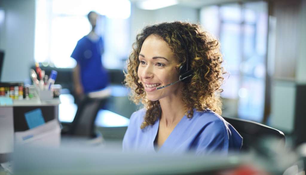 Smiling woman in scrubs with a headset at a desk.