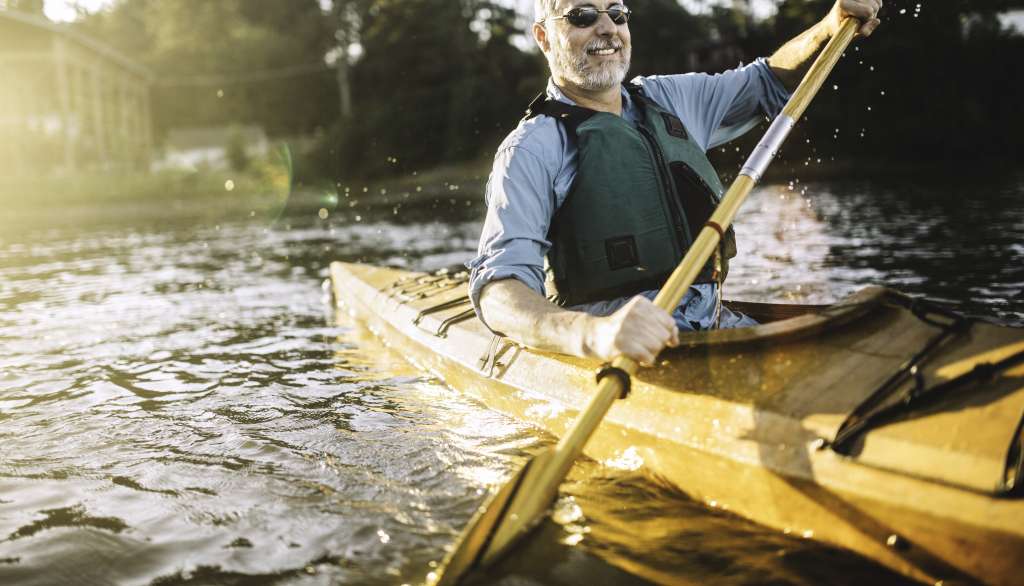 Man kayaking on a sunlit lake, smiling, wearing sunglasses and a life jacket.