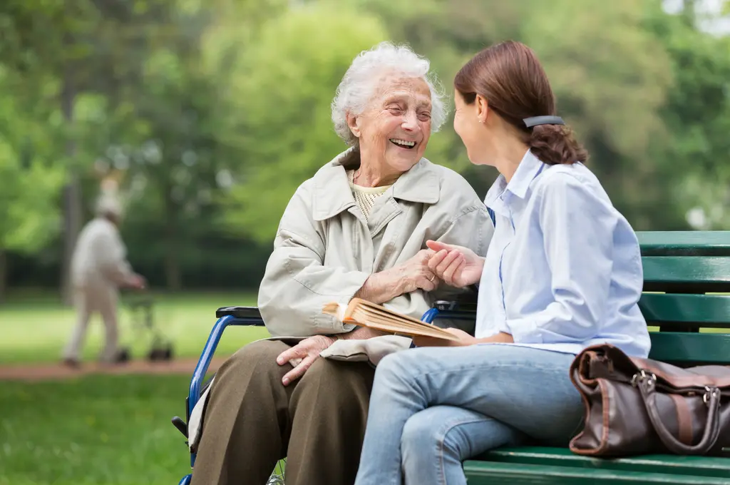 Elderly woman and young woman smiling, sitting on a park bench, holding hands.