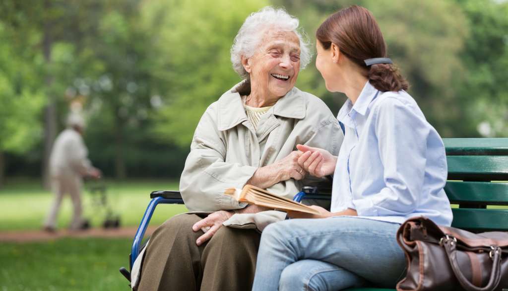 Elderly woman and young woman smiling, sitting on a park bench, holding hands.