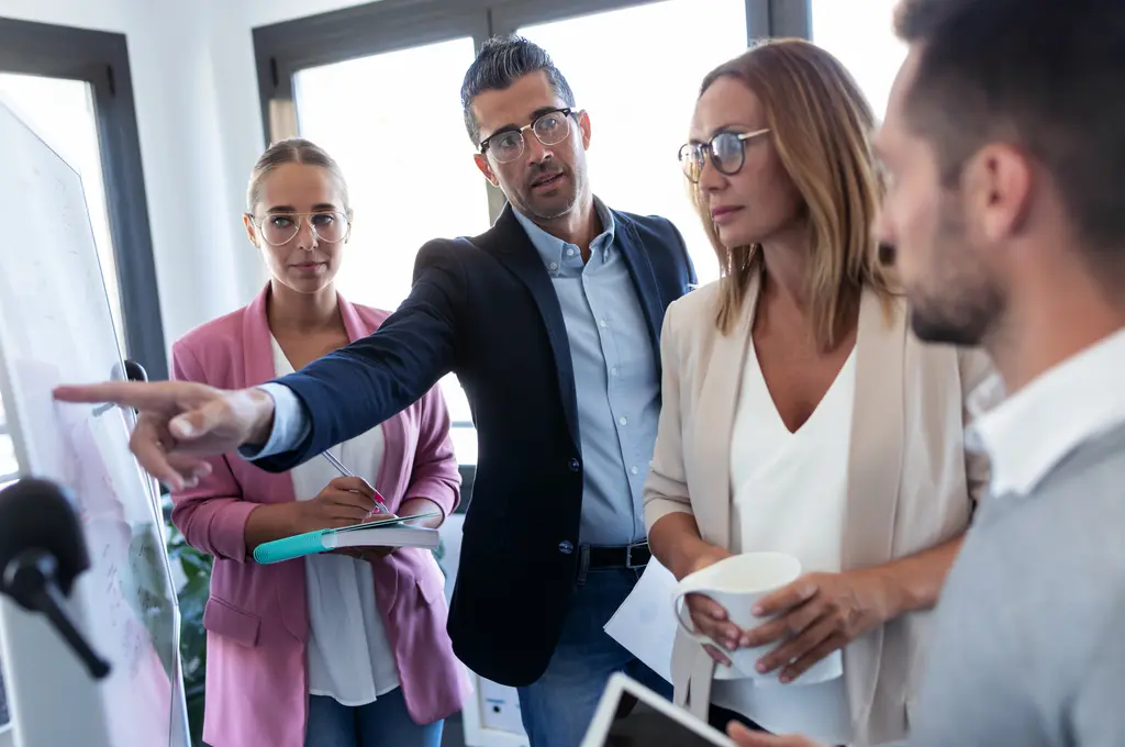 Group of colleagues in a meeting, one pointing at a whiteboard.