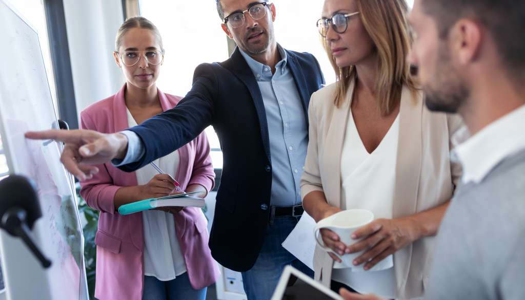 Group of colleagues in a meeting, one pointing at a whiteboard.