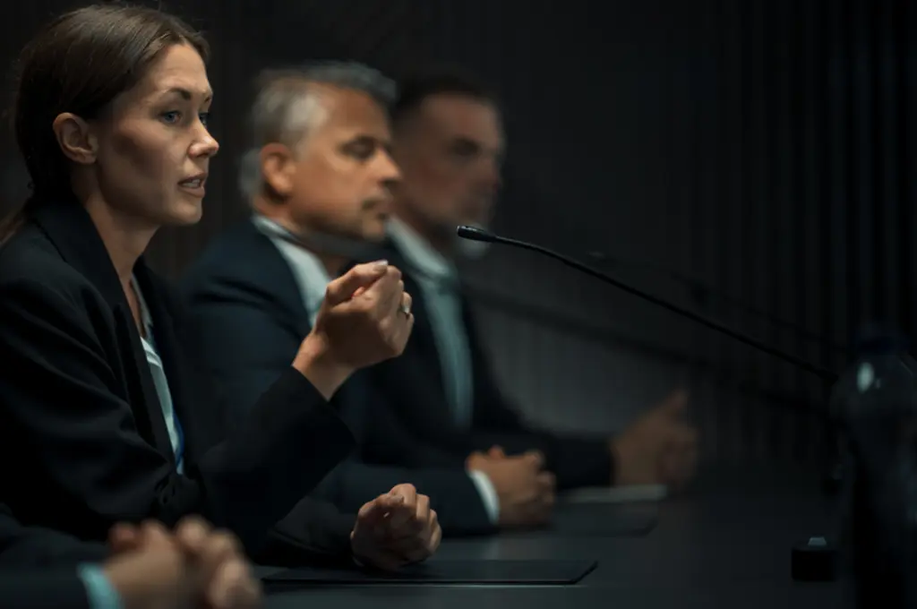 Women and men in suits at a conference table, one speaking into a microphone.