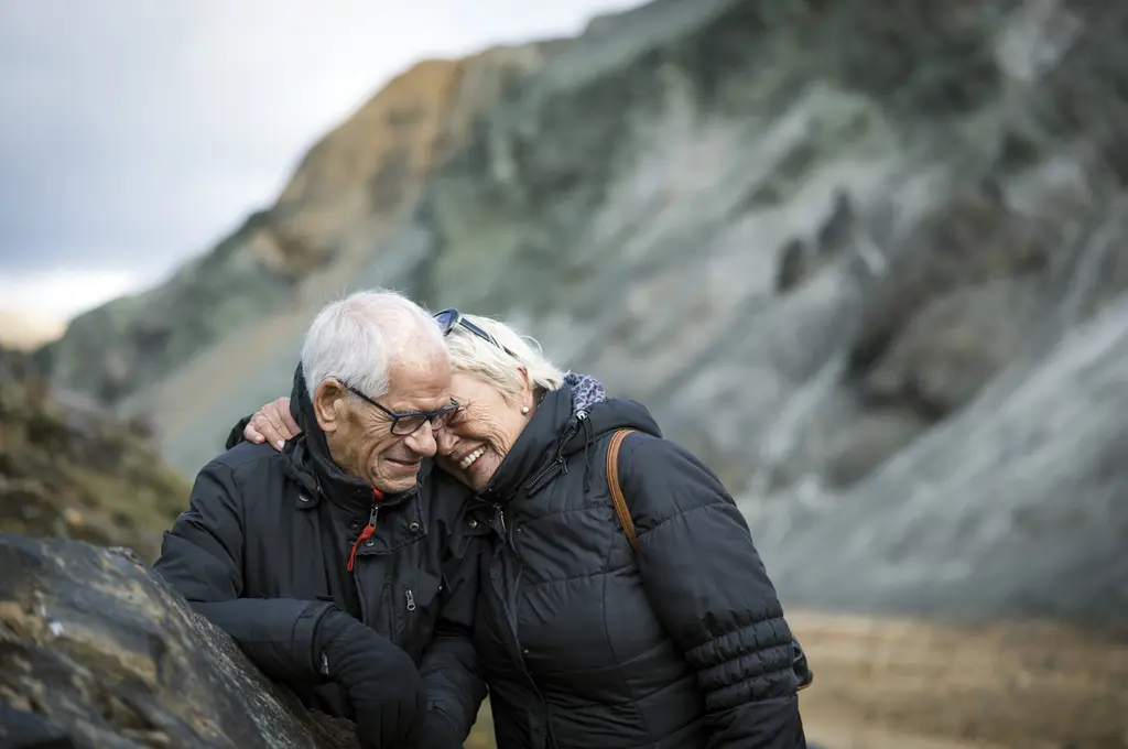 Elderly couple smiling, embracing outdoors in front of a rocky landscape.
