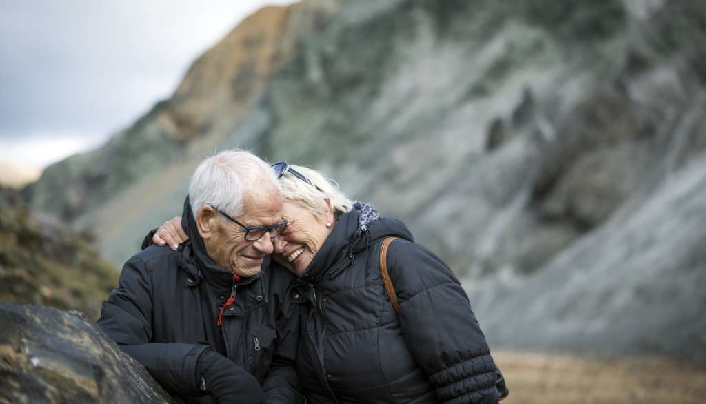 Elderly couple smiling, embracing outdoors in front of a rocky landscape.