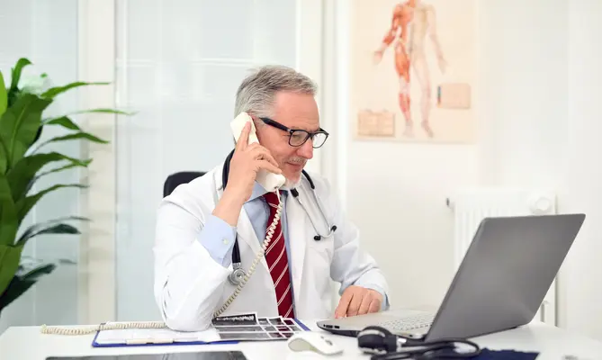 Male doctor talking on phone at desk with laptop open