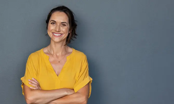 Smiling middle-aged woman with her arms crossed leaning against a grey wall.
