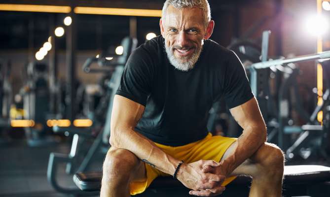 Man smiling, seated on a gym bench, wearing a black shirt and yellow shorts.