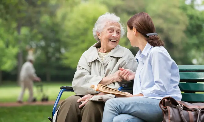 Elderly woman and young woman smiling, sitting on a park bench, holding hands.