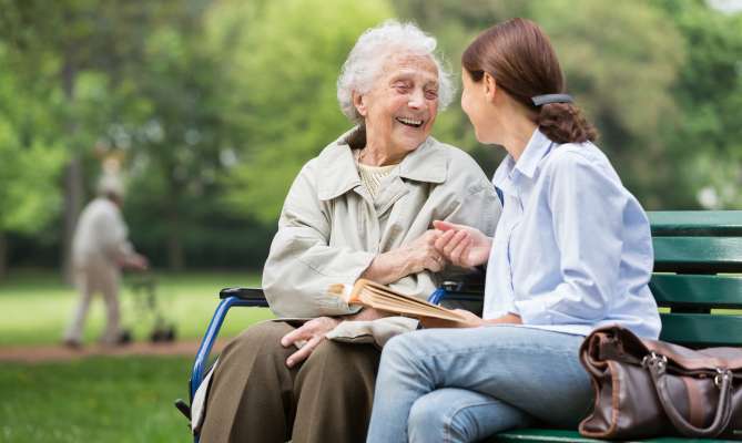 Elderly woman and young woman smiling, sitting on a park bench, holding hands.