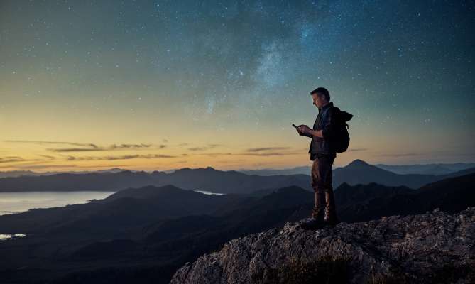 Person with backpack standing on a mountain at sunset, looking at phone. Stars in the sky.