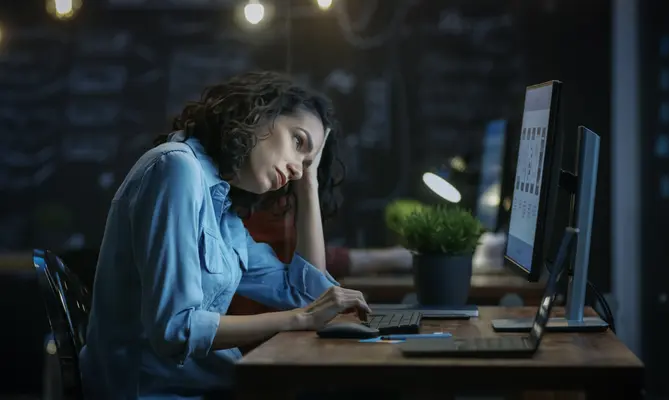 Woman in blue shirt, tired, working on a computer at night.