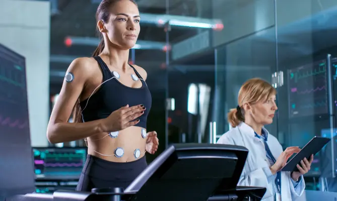 Woman on treadmill with sensors, doctor monitoring data on tablet.