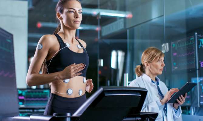 Woman on treadmill with sensors, doctor monitoring data on tablet.