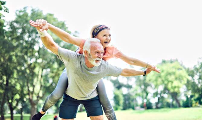 Elderly couple playing in a park, man gives woman a piggyback ride.