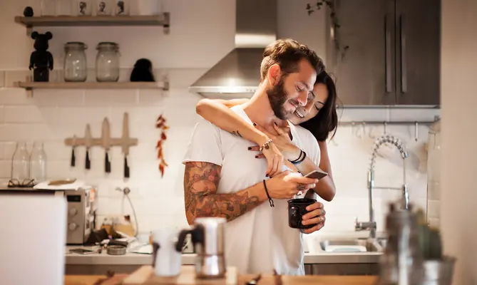 Couple embracing in a modern kitchen, smiling and looking at a phone.