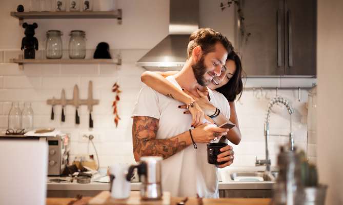 Couple embracing in a modern kitchen, smiling and looking at a phone.