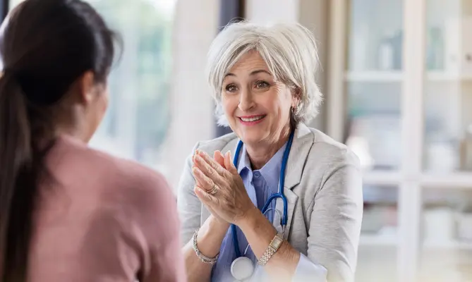 Smiling doctor in office speaks with patient, stethoscope around neck.