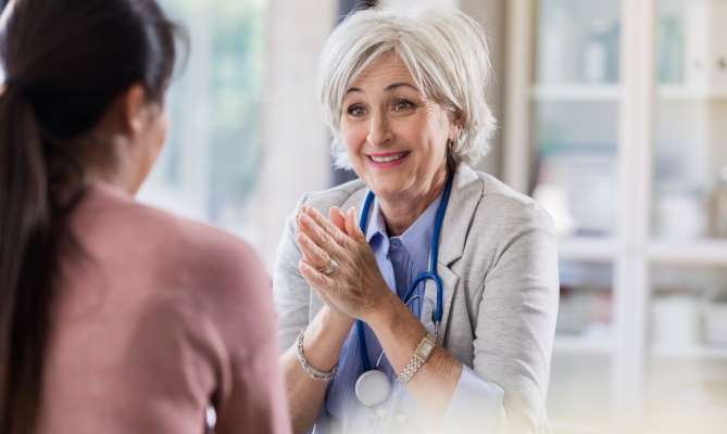 Smiling doctor in office speaks with patient, stethoscope around neck.
