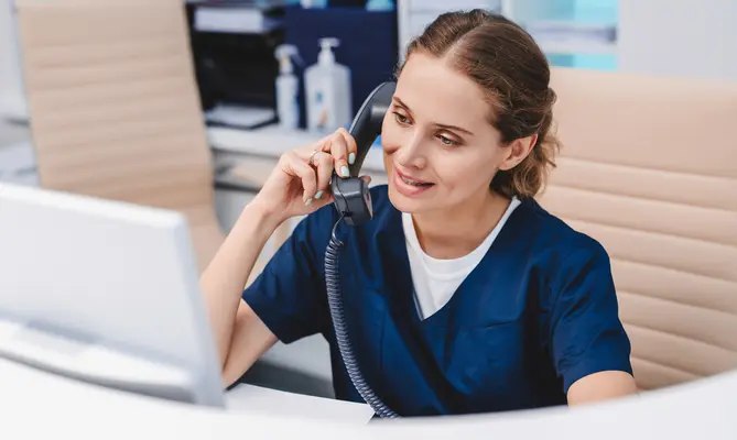 Nurse on phone at computer, wearing blue scrubs, smiling.