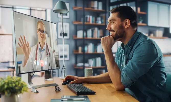 Man in a video call with a doctor on a computer, smiling in a modern home office.