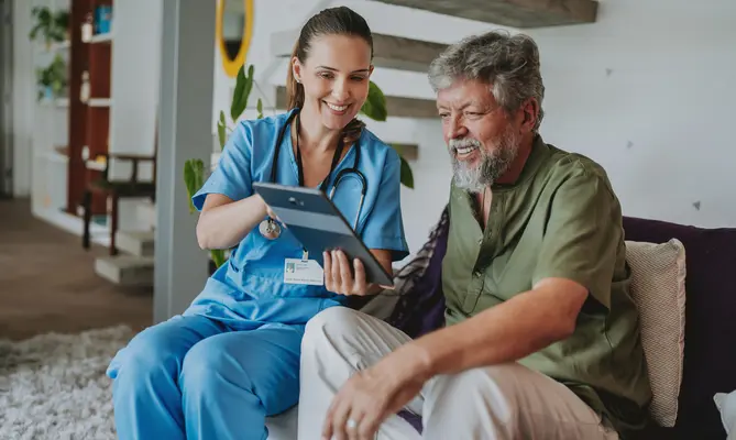 Nurse and elderly man smiling and looking at a tablet together in a living room.