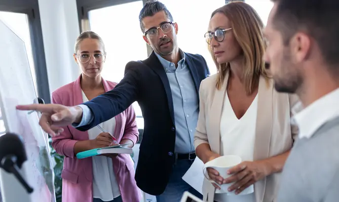 Group of colleagues in a meeting, one pointing at a whiteboard.