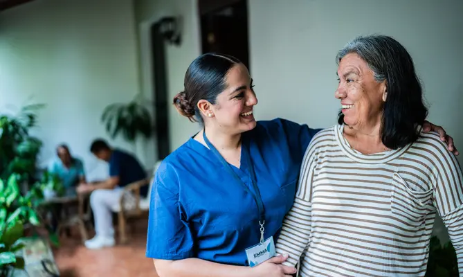 Caregiver and elderly woman smiling, standing in a bright room with plants.