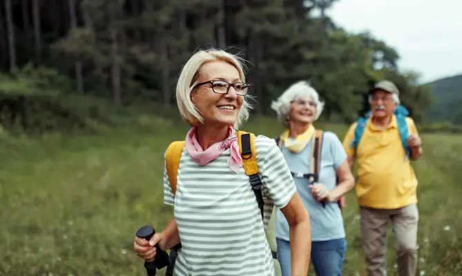 Elderly hikers smiling and walking through a grassy field.