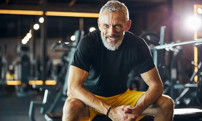 Man smiling, seated on a gym bench, wearing a black shirt and yellow shorts.