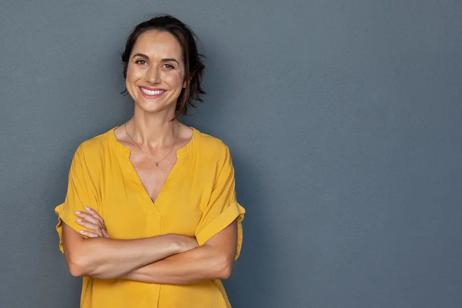 Smiling middle-aged woman with her arms crossed leaning against a grey wall.