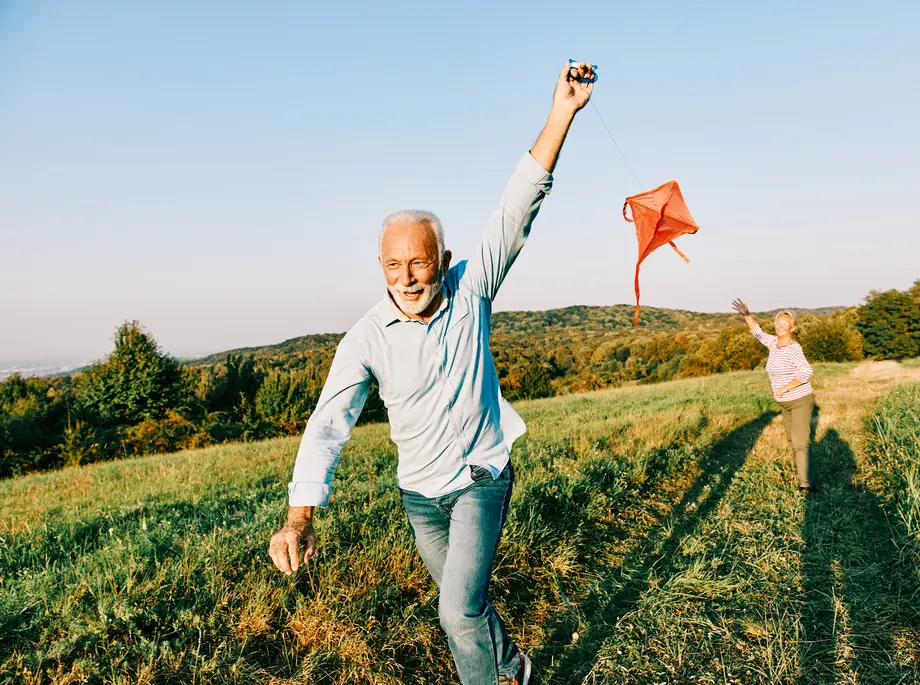 Happy middle-aged couple running and flying a red kite in an open field