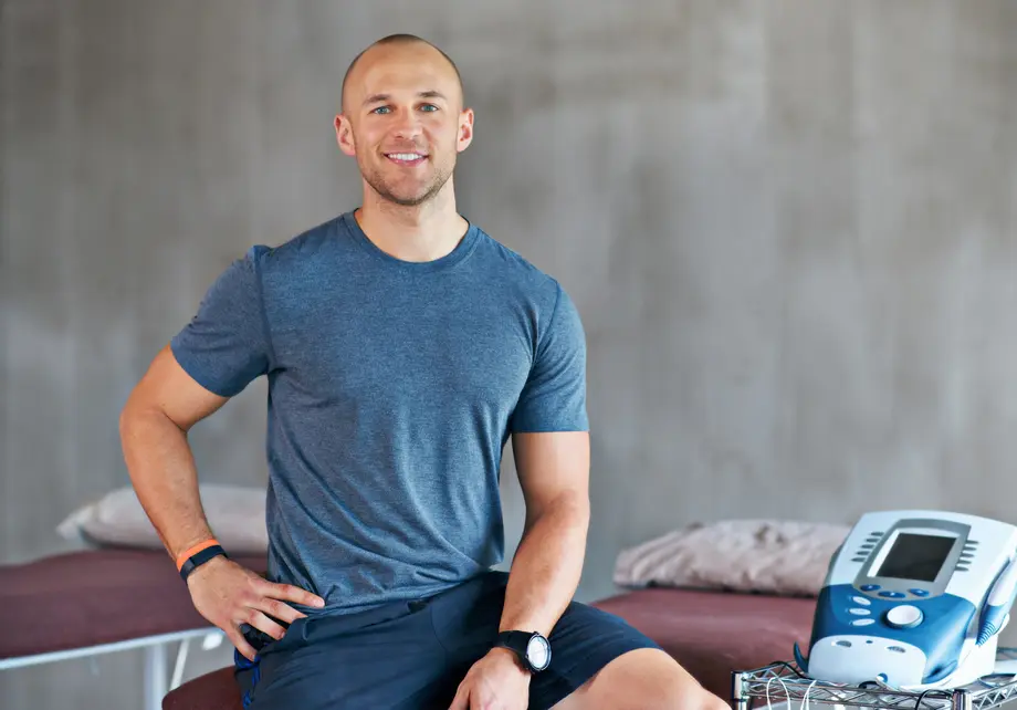 Man in a gym, smiling, sitting beside medical equipment.