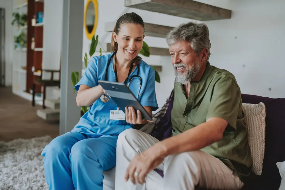Nurse and elderly man smiling and looking at a tablet together in a living room.