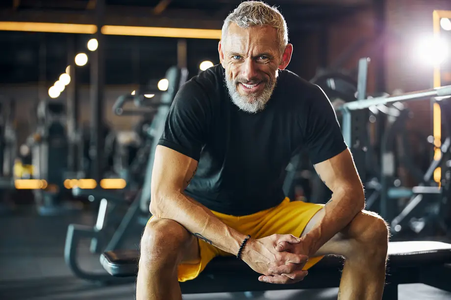 Man smiling, seated on a gym bench, wearing a black shirt and yellow shorts.
