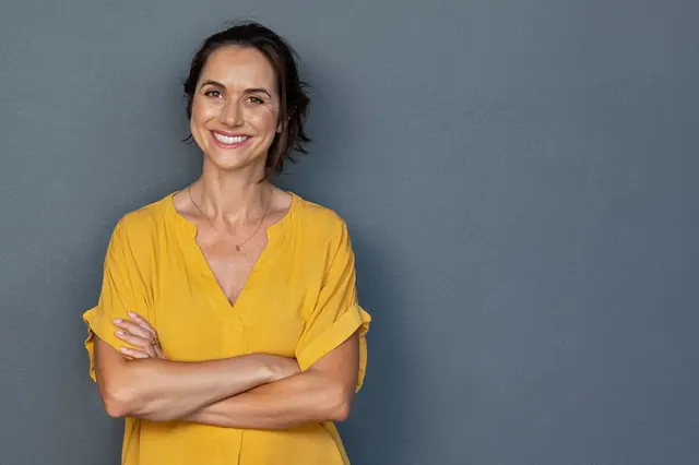 Smiling middle-aged woman with her arms crossed leaning against a grey wall.