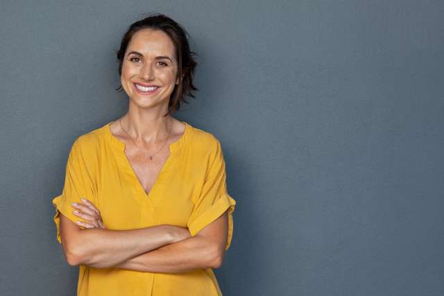 Smiling middle-aged woman with her arms crossed leaning against a grey wall.