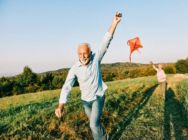 Happy middle-aged couple running and flying a red kite in an open field