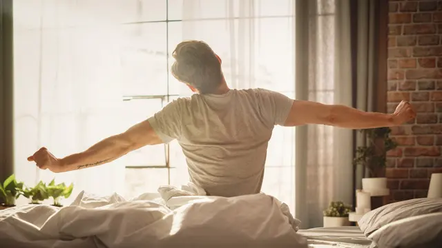 Man stretching in bed, morning light through window, relaxed atmosphere.