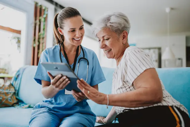 Nurse and elderly woman smiling, discussing information on a tablet at home.