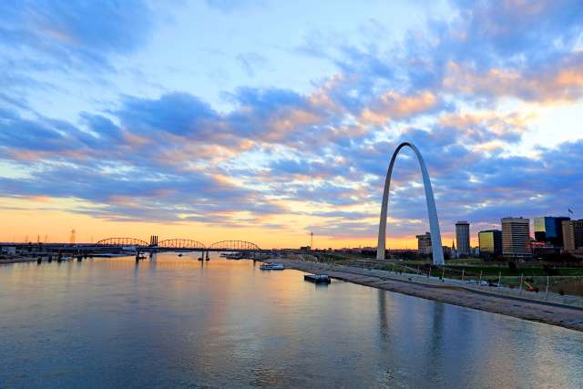 Gateway Arch with sunset-lit clouds reflecting on a river.